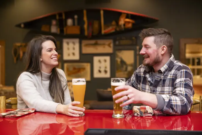 A man and woman sit at a wood-topped bar&#44; drinking glasses of beer. Rustic Adirondack decor is visible in the background.