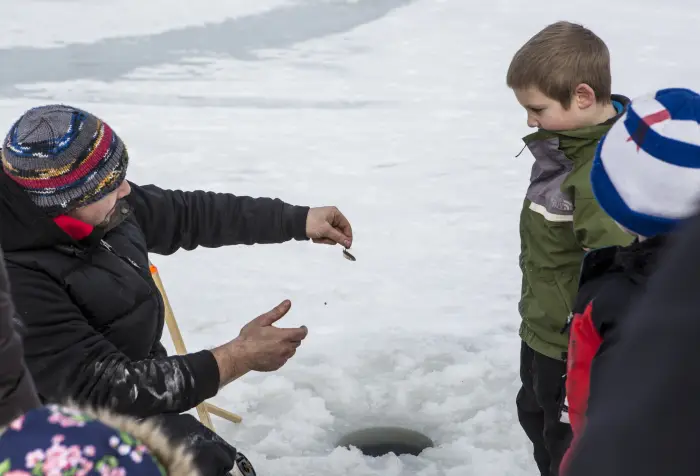 A man shows a fishing lure to kids on the ice next to an ice fishing hole.