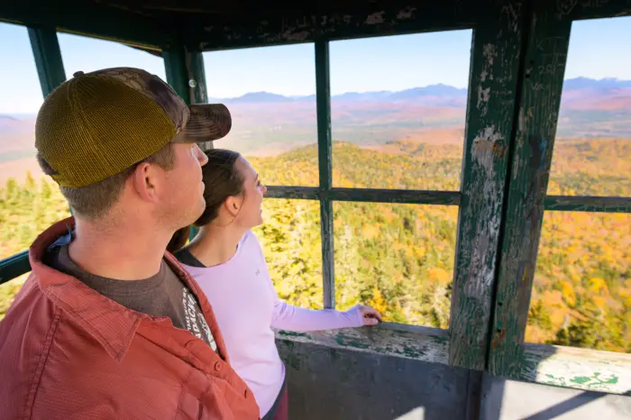 A couple stands in a fire tower cab with the forest below them in full fall colors.