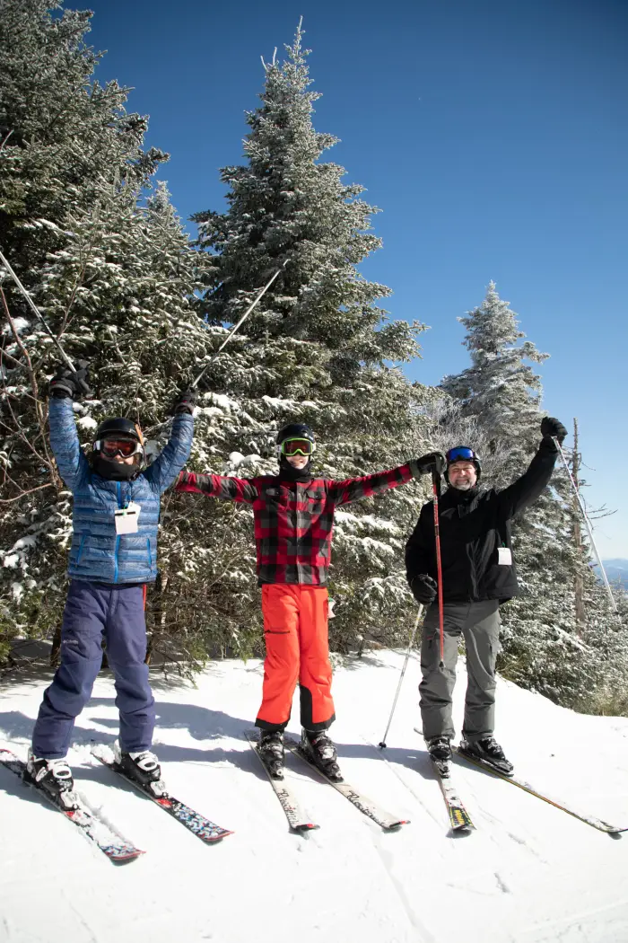 Three downhill skiers on a snowy slope raise their arms and smile