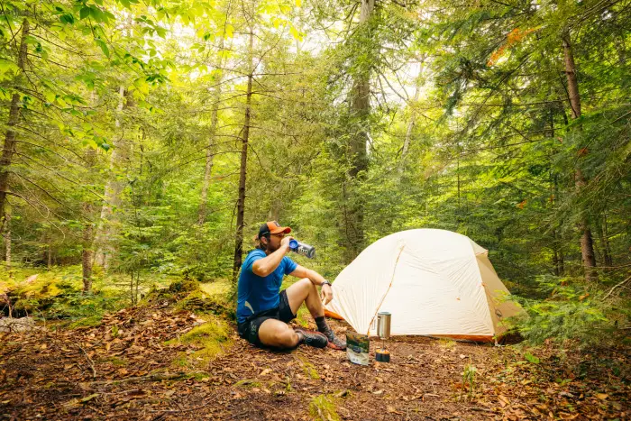 A man sits on the ground next to a tent in a forest.