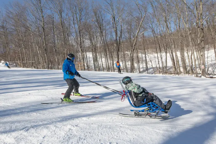 A skier in a blue goes gives a lesson to someone in a bucket style bi-ski.