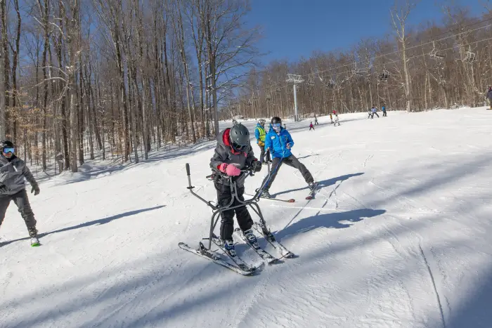 Someone in a blue coat gives an adaptive&#44; para ski lesson to someone who uses outriggers for support.