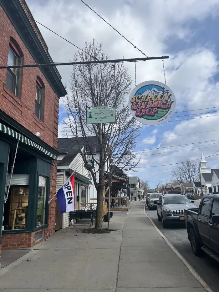 Schroon Lake Sandwich Shop sign hangs in front of a brick building on a small-town Main Street.