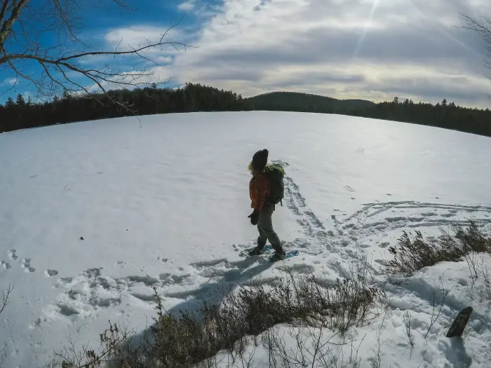 A woman in an orange sweater stands on a frozen lake in winter looking out over the wilderness.