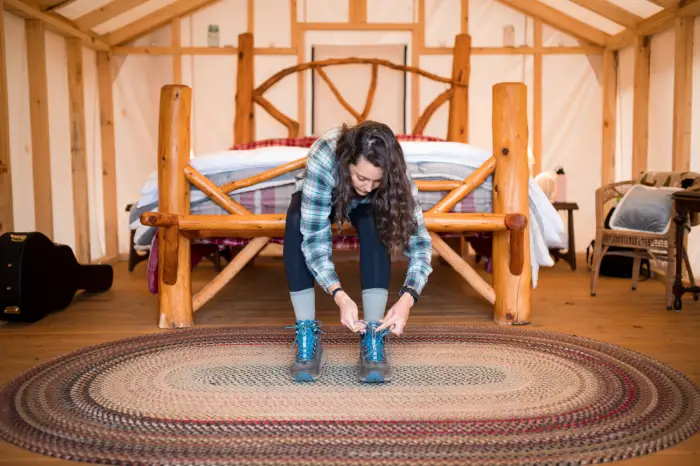 Woman puts on boots at the end of rustic looking bed in a glamping tent.