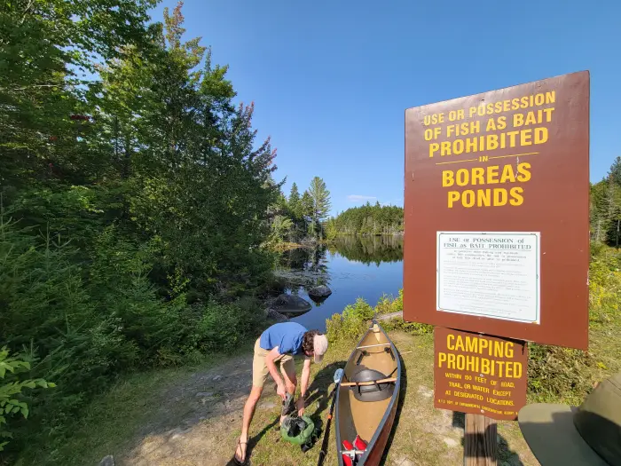 A paddler arranges gear in a canoe near a fishing regulation sign adjacent to water access