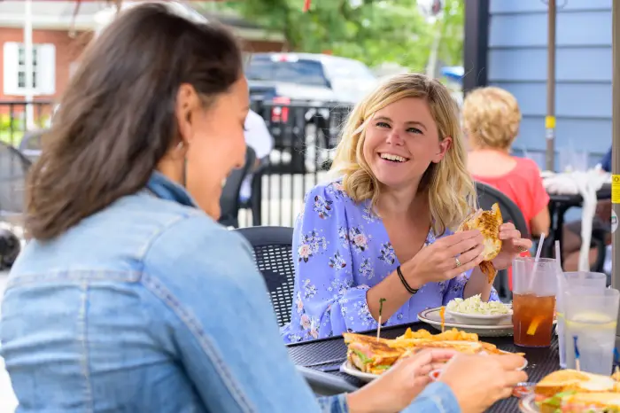 A woman laughs with her friend while eating a sandwich on a restaurant patio.
