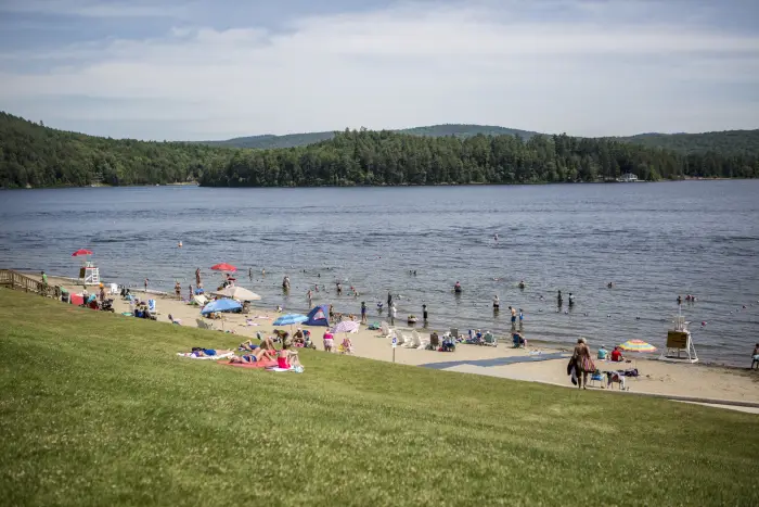 A wide view of people lounging and playing on a sandy beach&#44; while others swim in a broad&#44; blue lake.